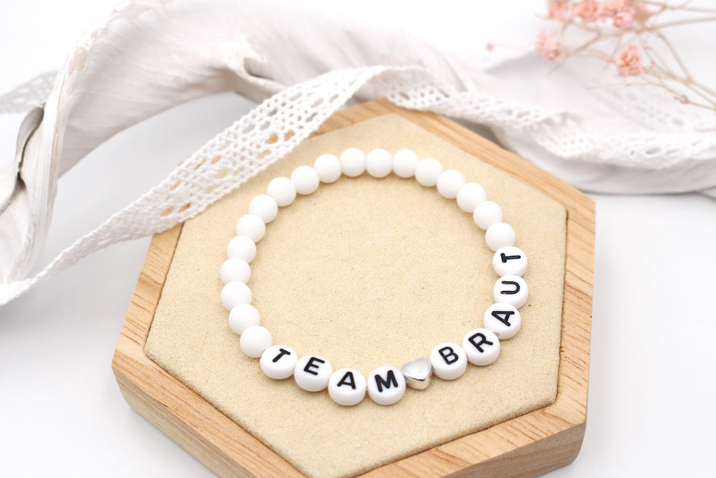 weiße Perlenarmbänder mit Team Braut und silberfarbenem Herzen auf beigen Holzbrettchen